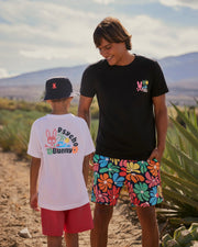 Two people stand outdoors on a sunny day, both wearing bright Psycho Bunny clothing. One, an adult in a black shirt and colorful MENS MENTZ SWIM TRUNK - B6W320B2SW by Psycho Bunny, faces and smiles at the other, a child in a white T-shirt and red shorts. There are plants and mountains in the background.
