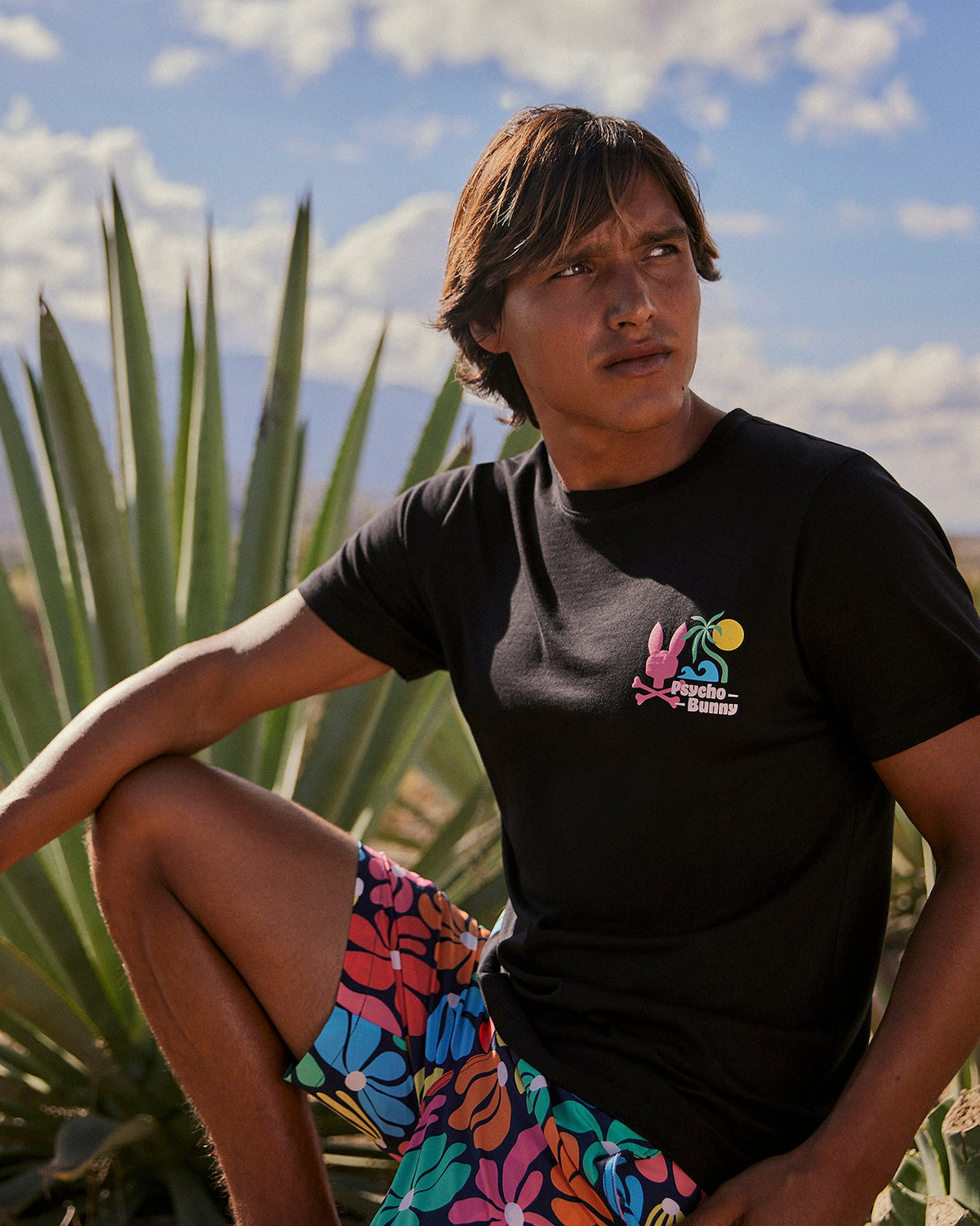 A person with shoulder-length hair sits outdoors in front of a large, spiky plant. They are wearing a black t-shirt with a colorful graphic on the chest and vibrant floral-pattern **Psycho Bunny MENS MENTZ SWIM TRUNK - B6W320B2SW**. The sky is partly cloudy with mountains in the background.