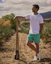 A man in a white t-shirt and aqua geometric-printed Psycho Bunny swim trunks leans on a shovel in a desert landscape, looking contemplatively to the side.