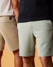 Two men standing side by side, focusing on their lower torsos. One wears a pair of Psycho Bunny MENS SHIRO KNIT HONEYCOMB SHORT in light beige, the other in light green; both shorts are in a casual style with a plain background.