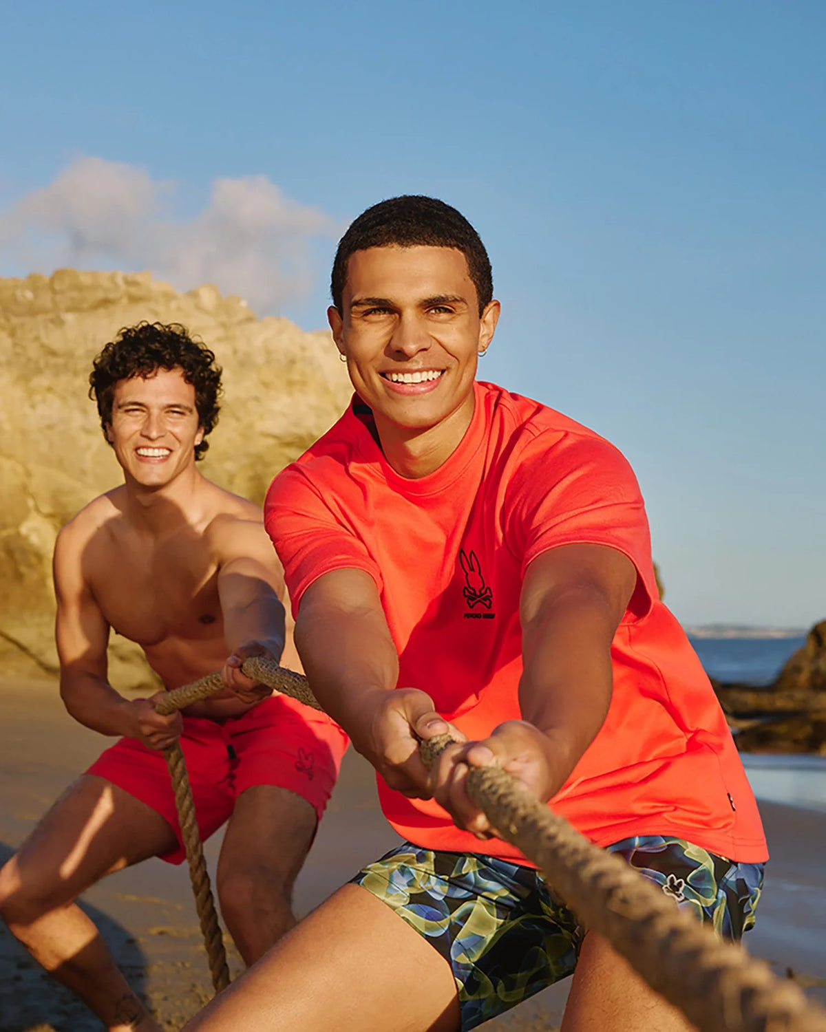 Two men are on a beach, engaged in a tug-of-war game with a rope. Both are smiling and appear to be enjoying themselves. The man in front wears an orange shirt made of heavyweight Pima cotton and patterned shorts—he's sporting the Psycho Bunny MENS BARRETT RELAXED FIT TEE - B6U518C200—while the man behind is shirtless and wears red shorts. The sky is clear and sunny.