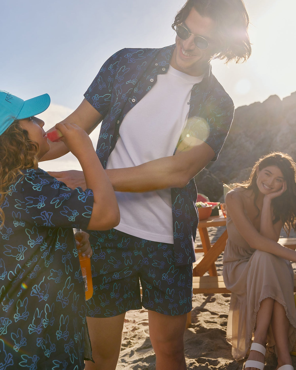 A man and a child wearing matching blue outfits with a bunny pattern enjoy a sunny day at the beach. The man, in Psycho Bunny MENS SHELDON ALL OVER PRINT SWIM TRUNK - B6W588C200, smiles while helping the child with a popsicle. A woman in a light dress sits on a picnic bench in the background, smiling toward them.