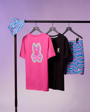 A clothing rack features a colorful bucket hat with a wave-like pattern, a pink t-shirt with a bunny skull graphic on the back, a black t-shirt with a small bunny skull on the front, and the Psycho Bunny MENS CLARKSON LIGHTWEIGHT SWIM TRUNK - B6W120B2SW. The background is a soft purple.