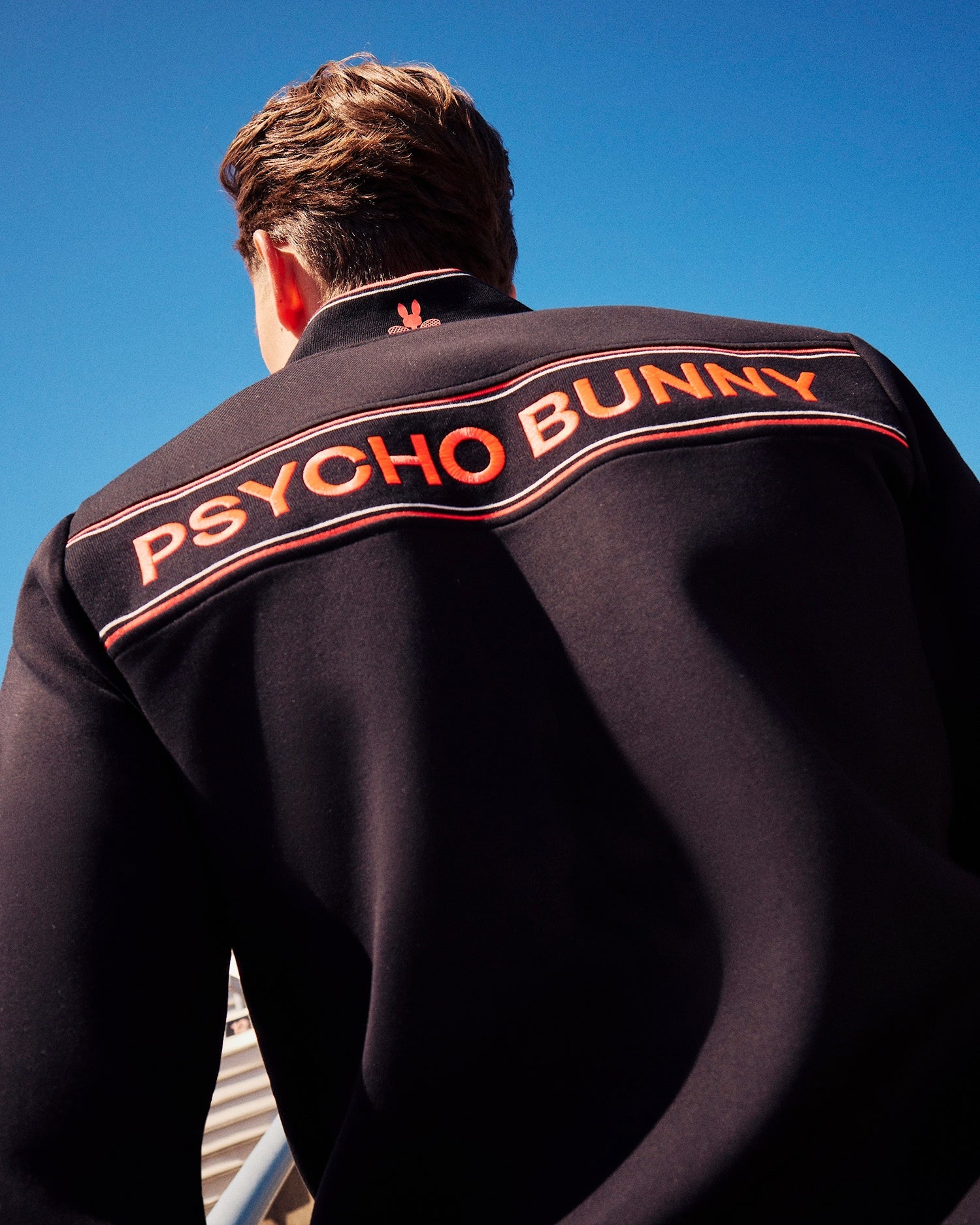A person with short brown hair is photographed from behind, wearing a MENS COURTSIDE TRACK JACKET - B6J692C200 featuring "PSYCHO BUNNY" in bold orange letters across the upper back. The dark jacket, crafted from stretch cotton-blend jersey, stands out against the clear blue sky.