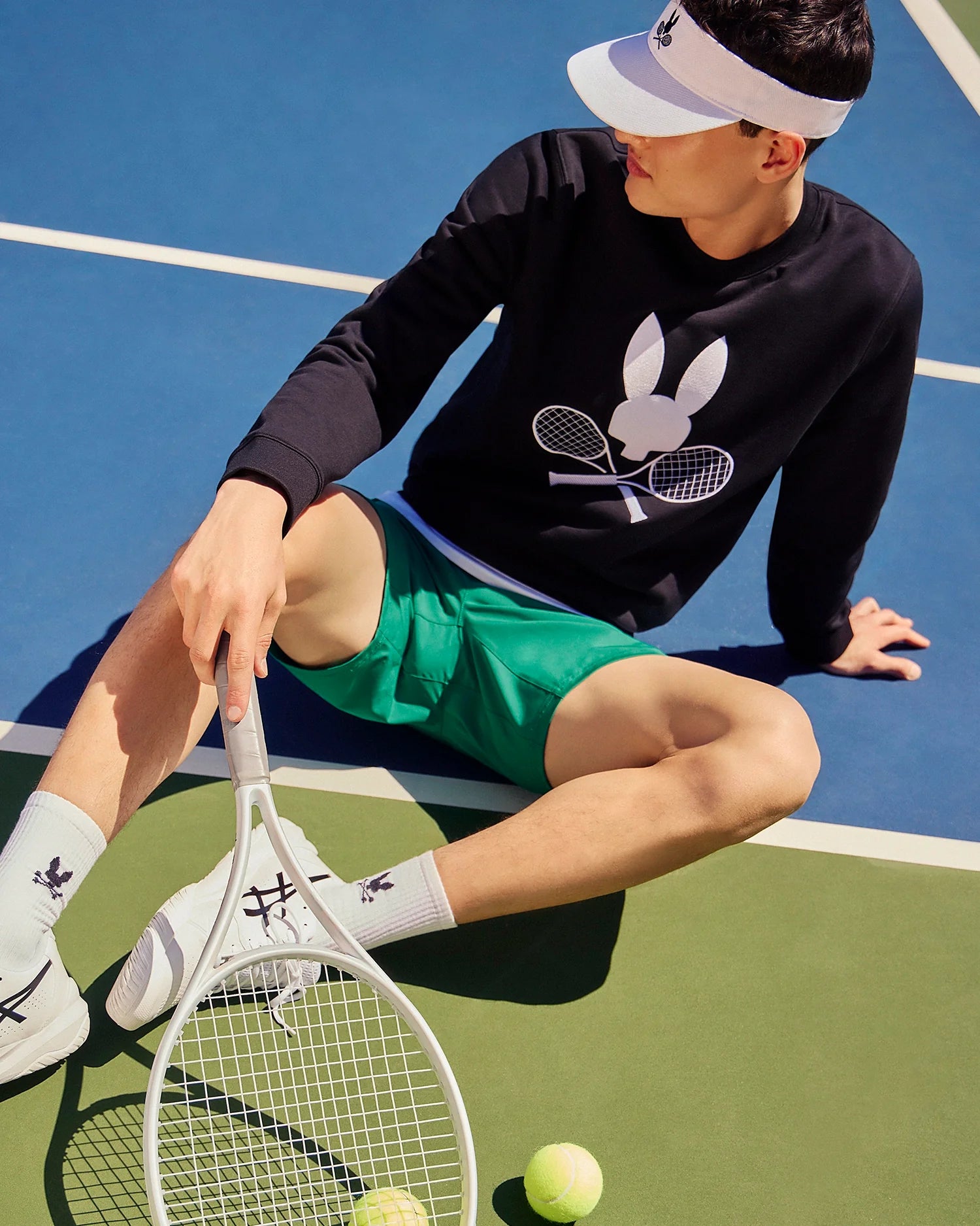 A person sits on the tennis court, wearing a white visor and a black Psycho Bunny MENS COURTSIDE SWEATSHIRT - B6S688C200 adorned with a bunny and crossed racquets. Complemented by green shorts and white socks, this ensemble adds flair while they hold a tennis racquet alongside the ball on the lush green court.