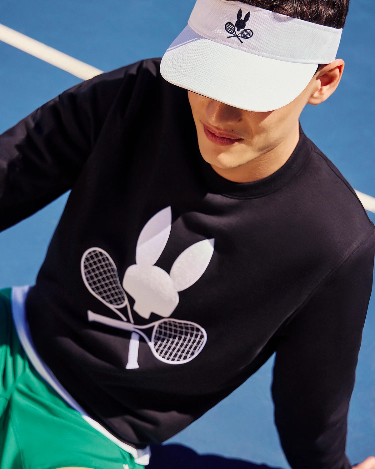 The individual is seated on a blue tennis court, wearing a white visor emblazoned with Psycho Bunny's logo and a black MENS COURTSIDE SWEATSHIRT - B6S688C200 from the same brand, showcasing the bunny with rackets motif. They finish off their look with green shorts made from a breathable cotton-blend fabric.