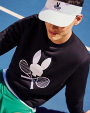 The individual is seated on a blue tennis court, wearing a white visor emblazoned with Psycho Bunny's logo and a black MENS COURTSIDE SWEATSHIRT - B6S688C200 from the same brand, showcasing the bunny with rackets motif. They finish off their look with green shorts made from a breathable cotton-blend fabric.