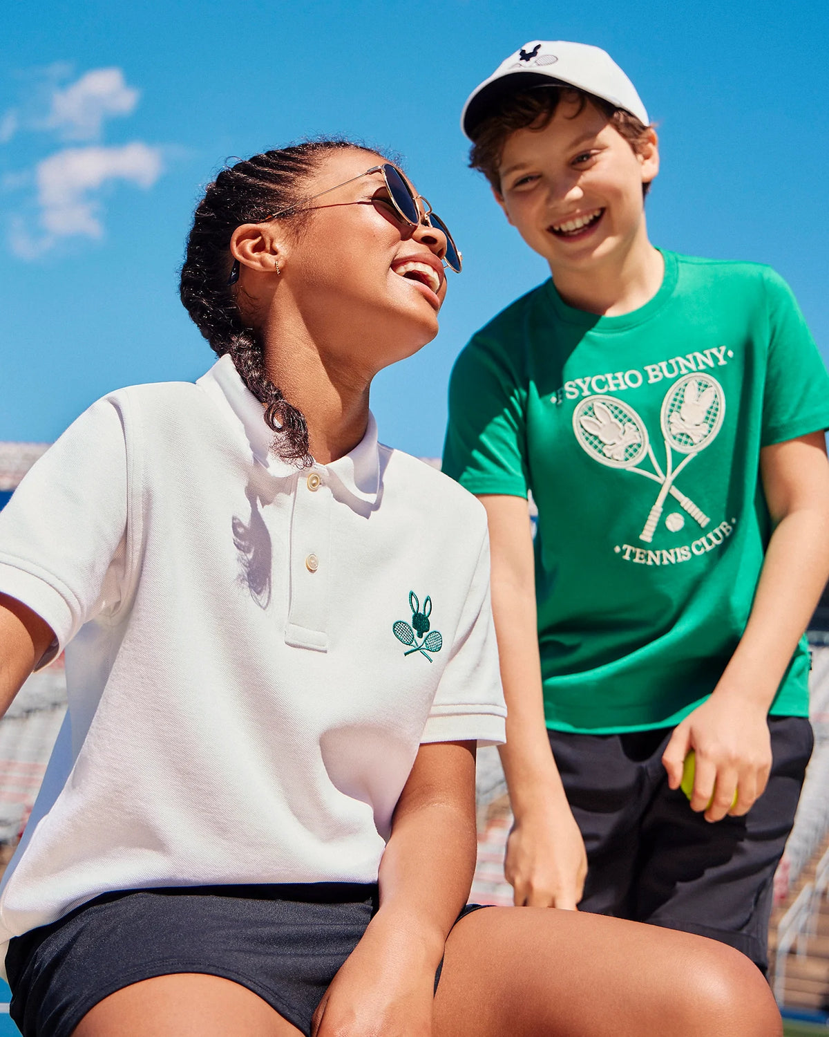 Two people are outdoors on a sunny day at a tennis court. One person, sitting and smiling upwards, wears a white polo shirt with the Psycho Bunny logo and sunglasses. The other person, standing and smiling, wears the green Kids' Courtside Graphic Tee by Psycho Bunny (product code B0U684C200) made of Pima cotton jersey and a cap.