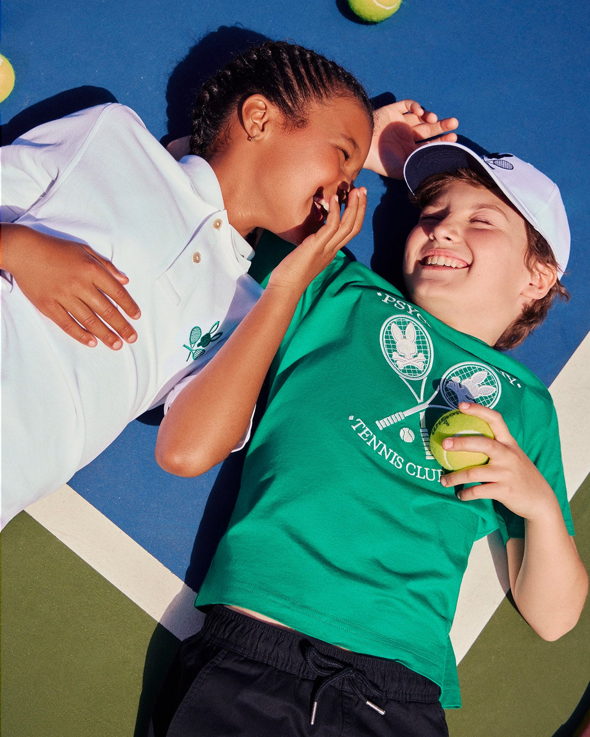 Two children lie on a tennis court, smiling and laughing. One child has their hand partially over their face while whispering to the other. There are tennis balls scattered around them. One wears a green shirt and white cap with the Psycho Bunny logo, while the other sports a white KIDS COURTSIDE GRAPHIC TEE by Psycho Bunny (B0U684C200).