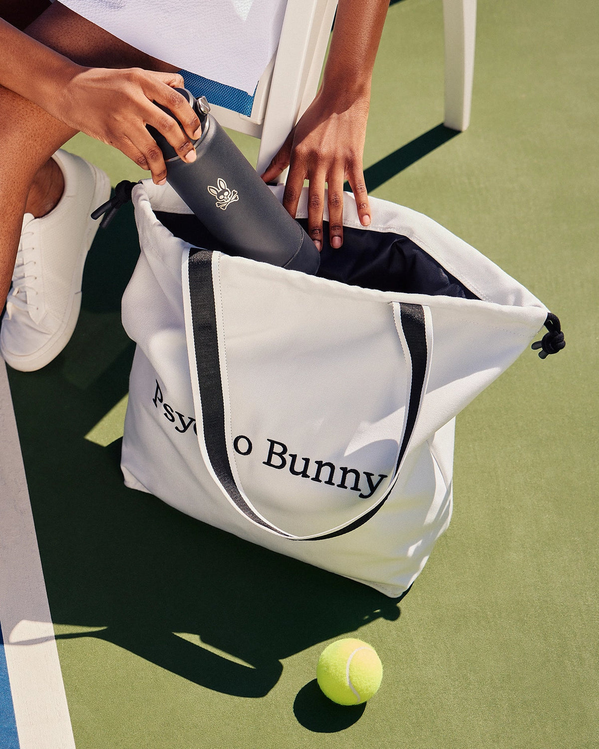 A person on a tennis court puts a black water bottle into a white Psycho Bunny TENNIS TOTE - B6A596C200. A yellow tennis ball is nearby, and the person wears white sneakers.