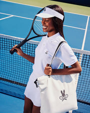 A person in a white polo, skirt, and visor with an embroidered tennis logo stands on a vivid blue tennis court, holding a Psycho Bunny TENNIS TOTE - B6A596C200. The net serves as the stylish backdrop for this ensemble.