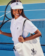 A woman in a white visor and polo shirt is on a tennis court, smiling with her racket and canvas Psycho Bunny TENNIS TOTE - B6A596C200. Both items showcase embroidered logos of rabbits and tennis rackets.