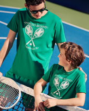 Two people, an adult and a child, enjoy their time on a tennis court. Both are clad in green t-shirts featuring the "PSYCHO BUNNY TENNIS CLUB" text and crossed racquets graphic. The adult wears sunglasses, and they share smiles while proudly showcasing the Psycho Bunny Kids Courtside Graphic Tee - B0U684C200, crafted from soft Pima cotton jersey.
