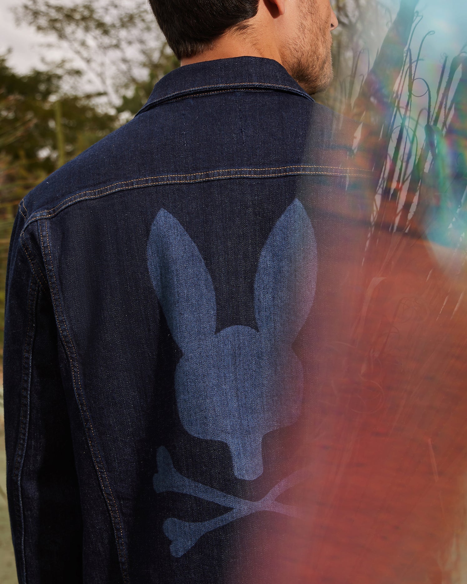 A man wearing the MENS PATHE DENIM JACKET - B6J741X1CO by Psycho Bunny, which features embroidered details of a bunny and crossbones on the back, stands outdoors. The scene includes blurred colorful elements in the foreground and trees in the background.