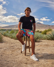 A person with long hair sits on a wooden stool in an outdoor setting with agave plants, mountains, and a blue sky in the background. They are wearing a black T-shirt with a colorful design on the chest, Psycho Bunny MENS MENTZ SWIM TRUNK - B6W320B2SW with vibrant floral patterns, and white sneakers.