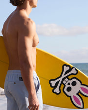 A young man stands on a beach holding a yellow surfboard with a skull and bunny graphic, looking toward the sea. He is shirtless and wearing light-colored Psycho Bunny MENS WILLIS STRETCH TENCEL shorts (B6R239Y1WB).