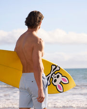 A young man with a surfboard, featuring a pink bunny design, stands facing the ocean. He is shirtless and wearing Psycho Bunny's MEN'S WILLIS STRETCH TENCEL SHORT - B6R239Y1WB, epitomizing a relaxed beach scene.