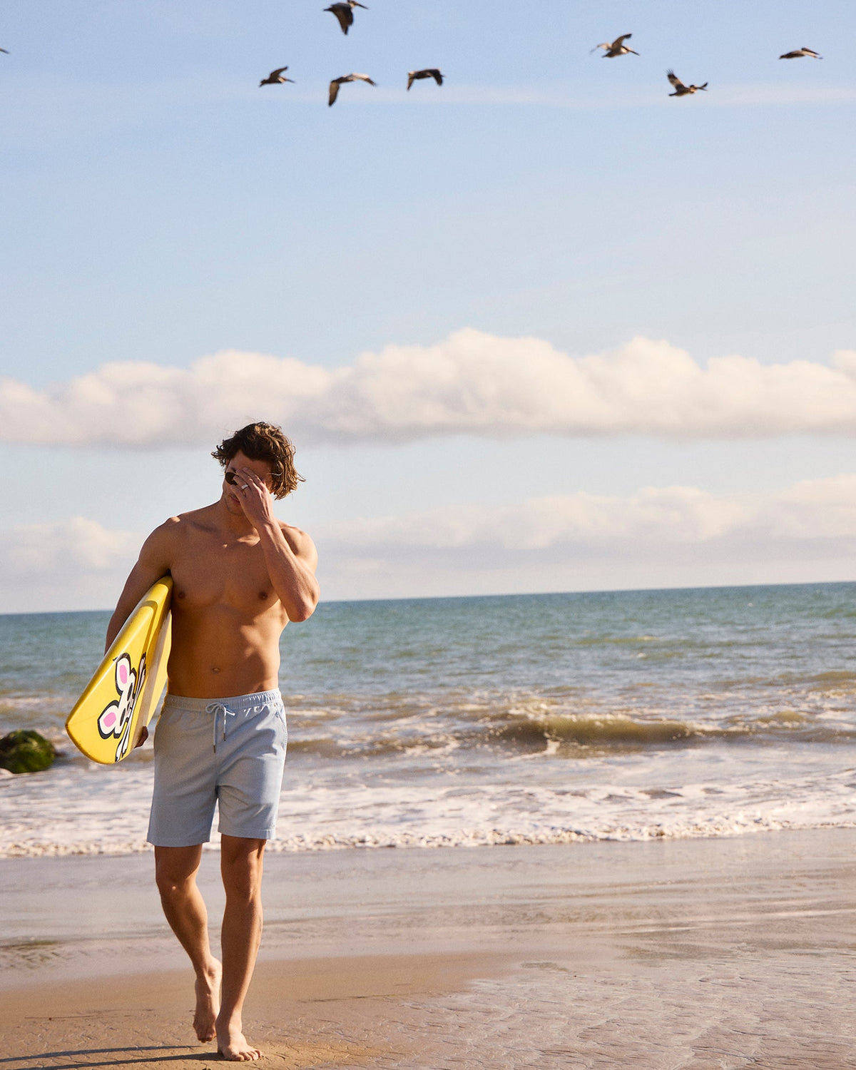 A man in Psycho Bunny Willis Stretch Tencel shorts walks on a sandy beach, holding a bright yellow skimboard. He gazes at the ocean while several birds fly overhead under a clear blue sky.