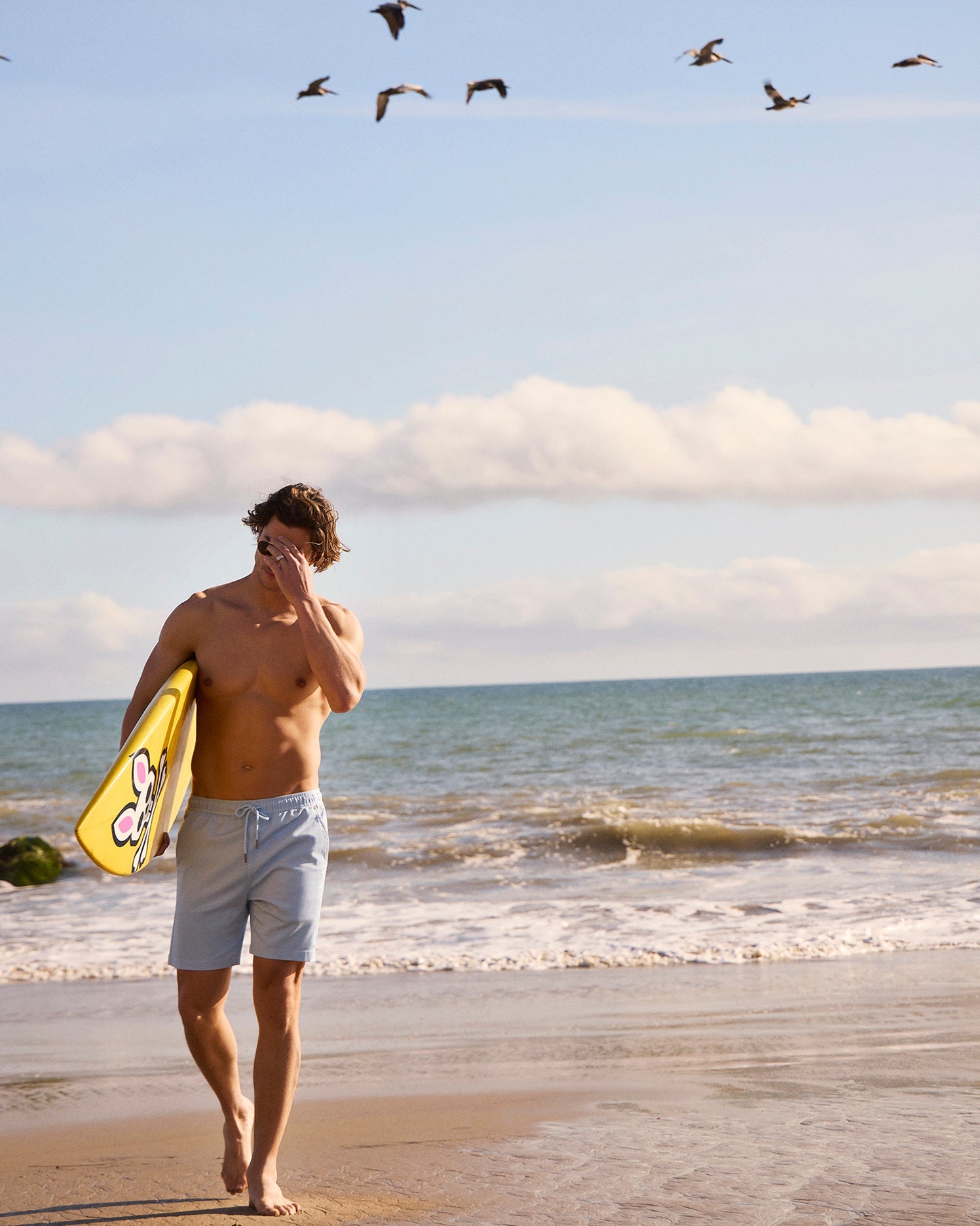 A man in Psycho Bunny Willis Stretch Tencel shorts walks on a sandy beach, holding a bright yellow skimboard. He gazes at the ocean while several birds fly overhead under a clear blue sky.