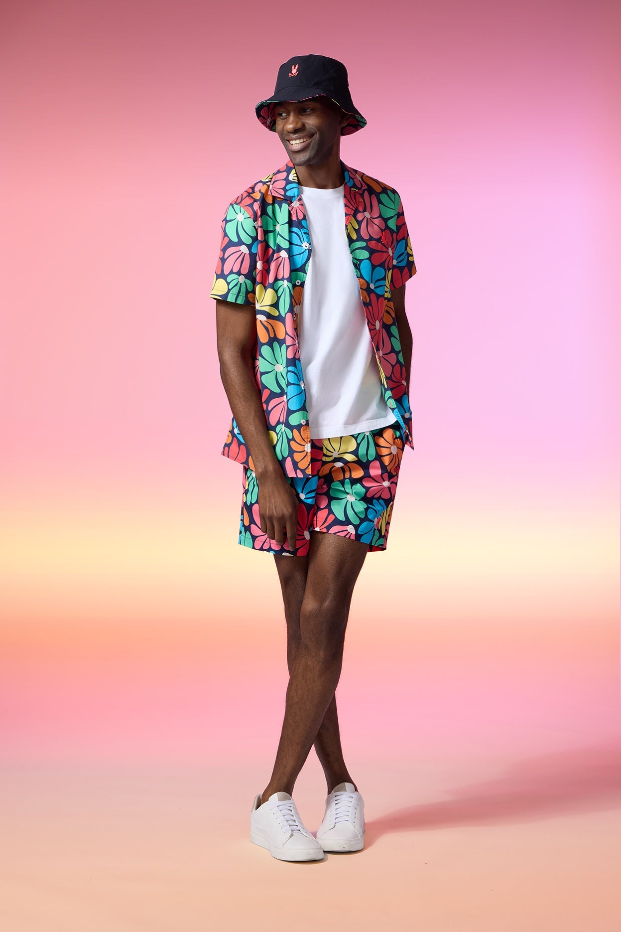 A person stands against a gradient pink and orange background, smiling. They wear a black bucket hat, a white t-shirt, and a matching colorful floral-pattern shirt and Psycho Bunny MENS MENTZ SWIM TRUNK - B6W320B2SW set, along with white sneakers. The outfit is bold and summery.