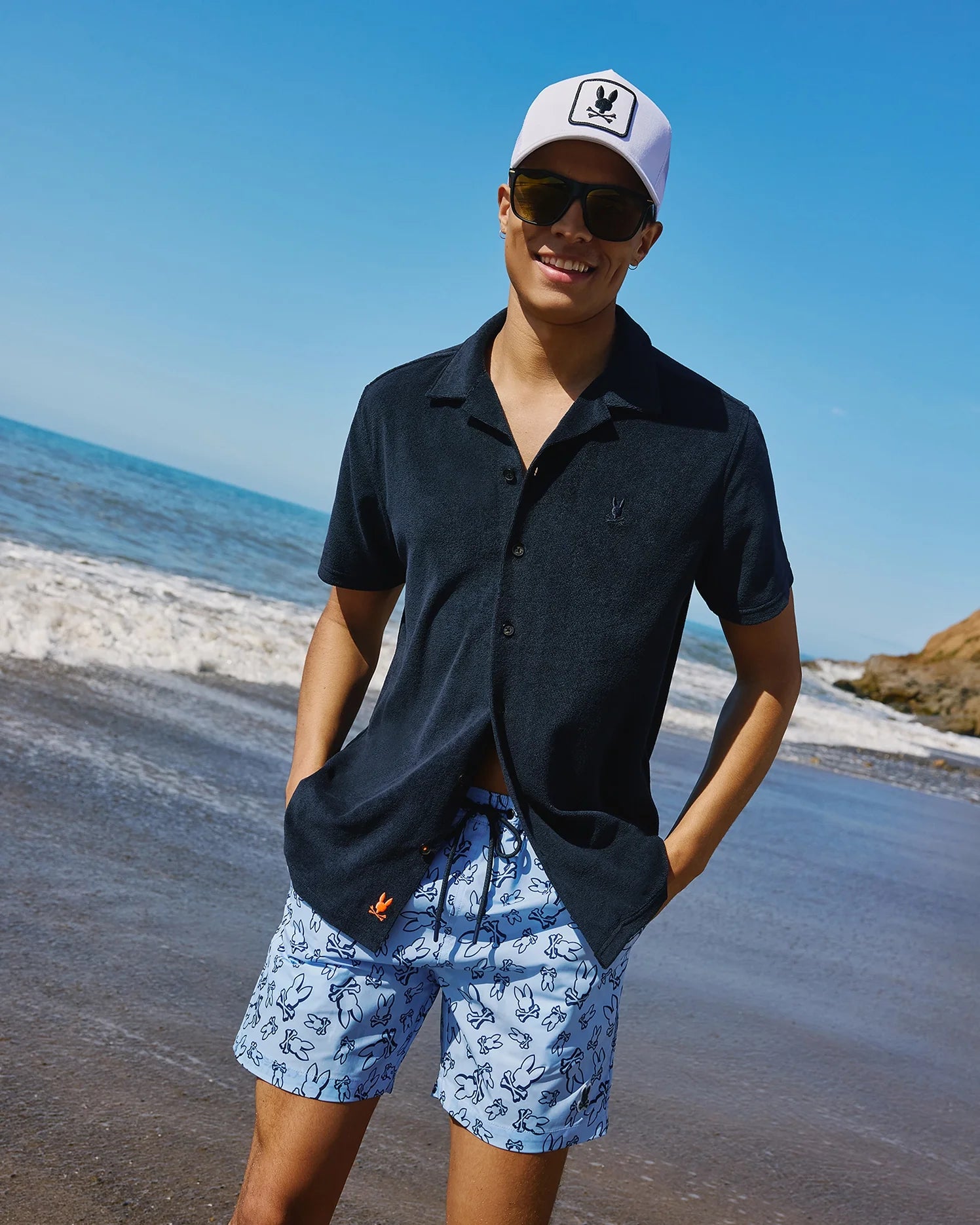 A smiling person stands on a sunny beach wearing a white baseball cap, sunglasses, a dark short-sleeve button-up shirt, and Psycho Bunny MENS RENDON PRINT SWIM TRUNK - B6W669C200 adorned with a bunny pattern. The ocean and part of a rocky cliff are visible in the background.