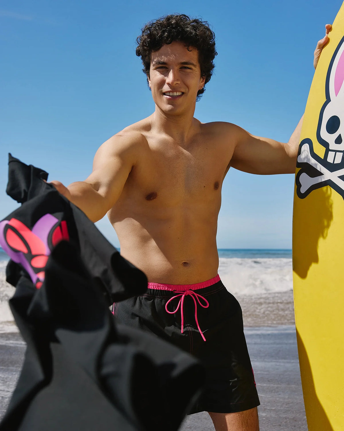 A man with curly hair smiles at the camera while standing on a beach. He is shirtless and wearing Psycho Bunny's MENS ROCKDALE LIGHTWEIGHT SWIM TRUNKS - B6W653C200, featuring pink drawstrings. In one hand, he holds a black shirt with a pink design, and in the other, he grips a yellow surfboard adorned with a skull and crossbones graphic. The ocean and clear blue sky stretch out behind him.

