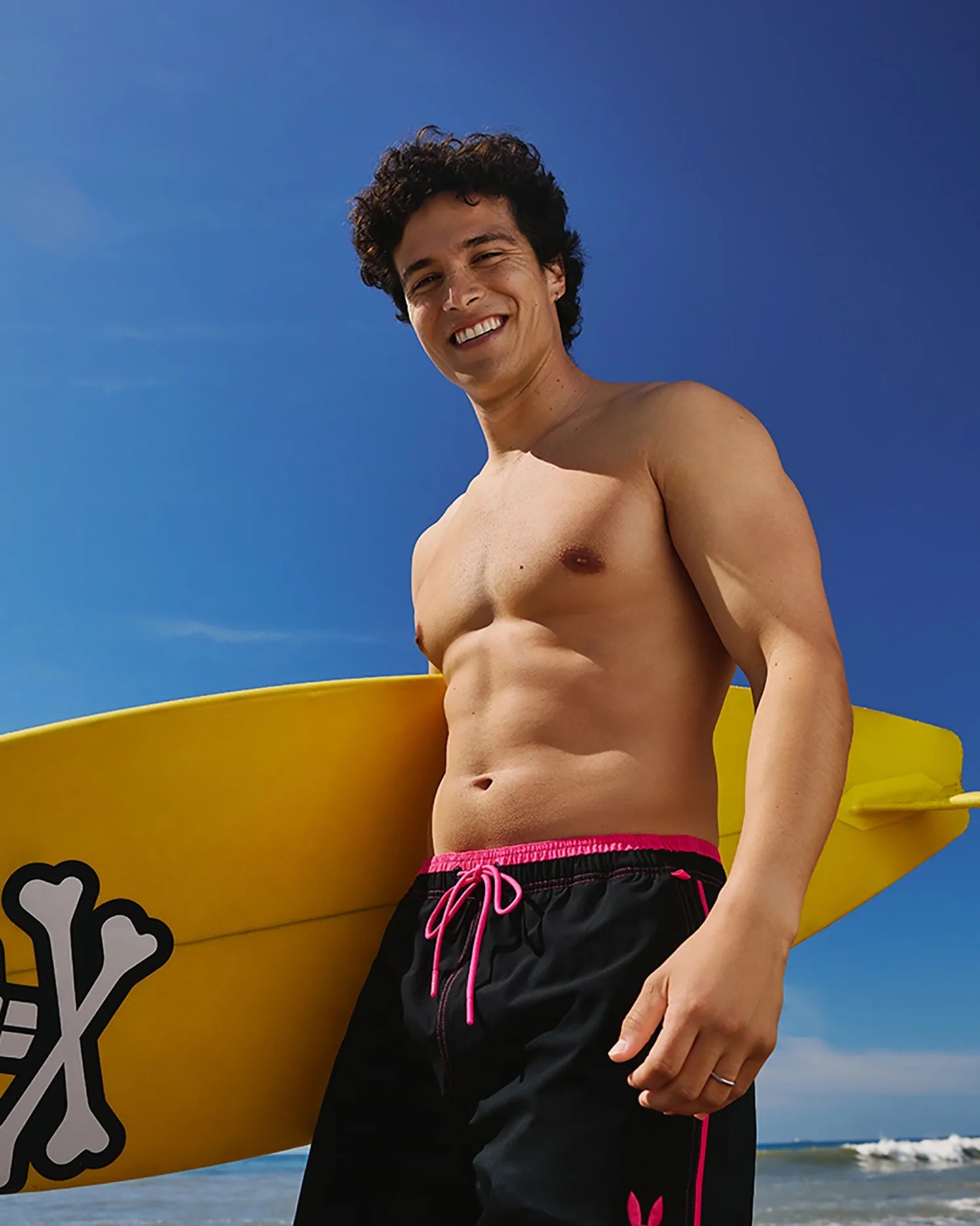 A smiling person stands on a beach, holding a yellow surfboard with a white logo. They have curly hair and are shirtless, wearing Psycho Bunny's MENS ROCKDALE LIGHTWEIGHT SWIM TRUNK - B6W653C200 black board shorts with pink drawstrings. The sky is clear and blue, and the ocean waves are visible in the background.
