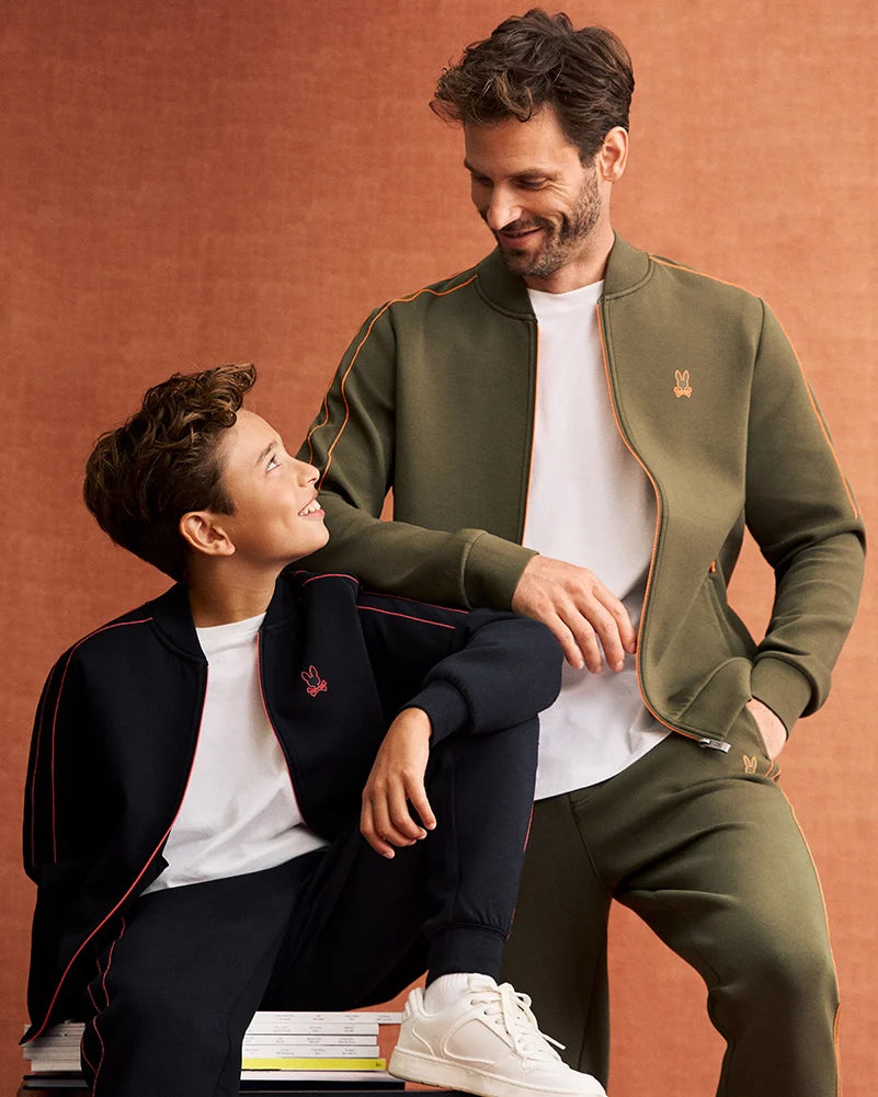 A man and a boy are sitting and smiling at each other. The man is wearing a matching olive green tracksuit with invisible zip pockets and a white shirt, while the boy is in a black MENS TRAVIS TRACK JACKET - B6J142D200 by Psycho Bunny with white sneakers. The background is a warm brown tone.