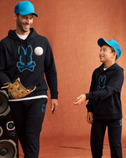 An adult and a child are standing together against an orange background. Both are wearing matching black outfits with blue Psycho Bunny KIDS MALTON TRUCKER CAPS - B0A797D200, hoodies displaying a rabbit and crossbones logo, and black pants. The adult is holding a baseball glove and ball, while smiling at the child.