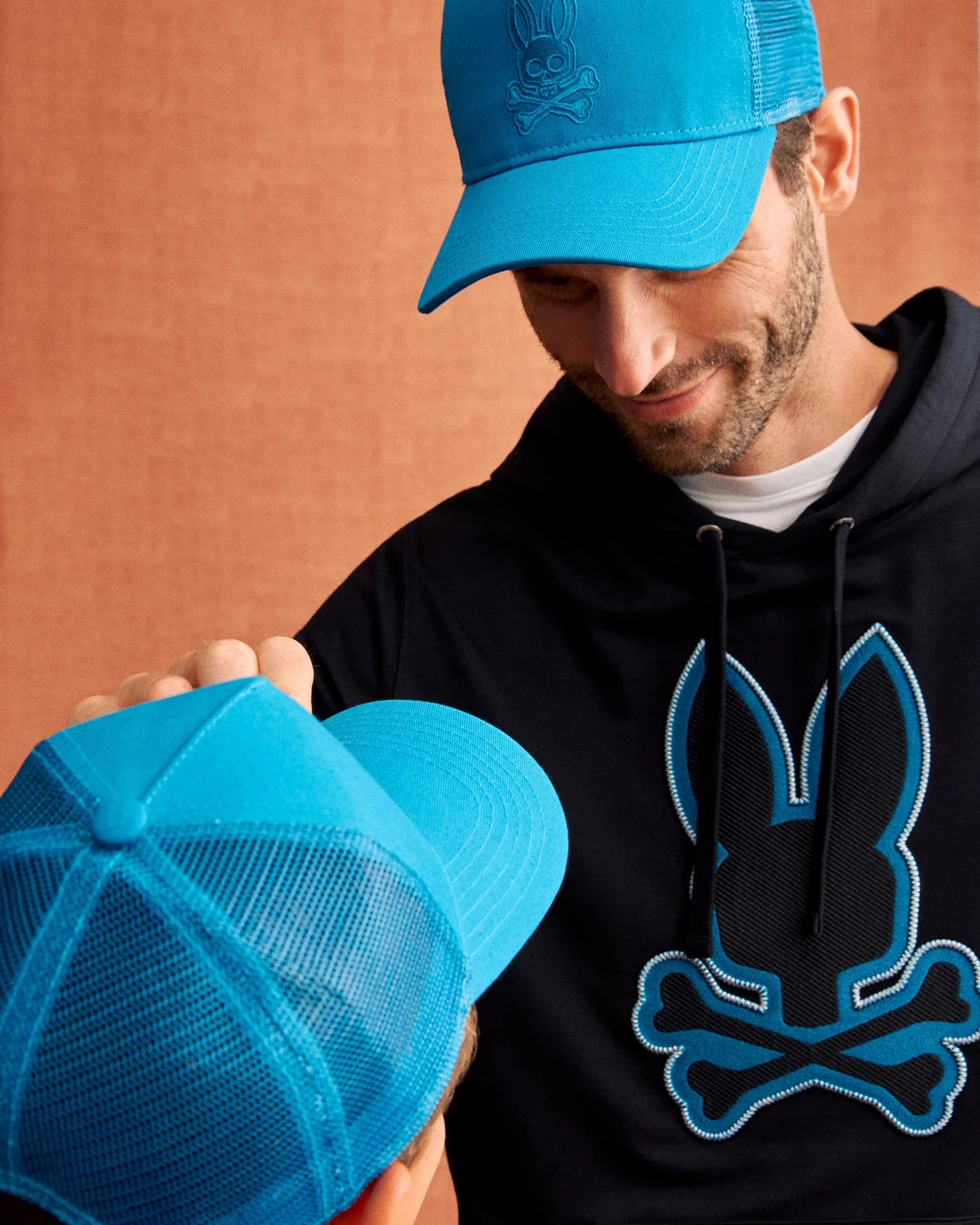 A man sports a bright blue trucker cap and a black hoodie with a blue outlined bunny skull logo from Psycho Bunny. He gazes down affectionately at a child in a matching KIDS MALTON TRUCKER CAP - B0A797D200. The background is a warm, brownish-orange color.