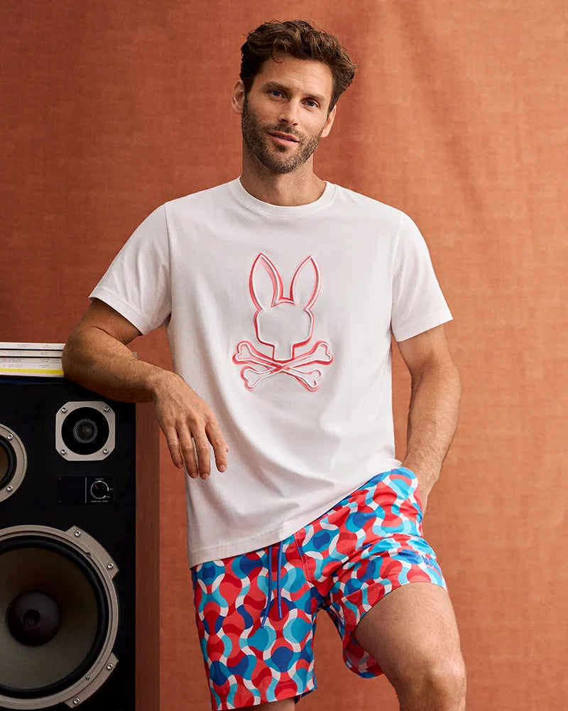 A man with short hair and a beard poses casually, leaning on a large speaker. He wears a white t-shirt adorned with a bunny skull design and MENS WALTER LIGHTWEIGHT PRINT SWIM TRUNK (B6W863D200) by Psycho Bunny, featuring a vibrant red, blue, and white pattern in 100% polyester. The background is a brownish fabric backdrop.