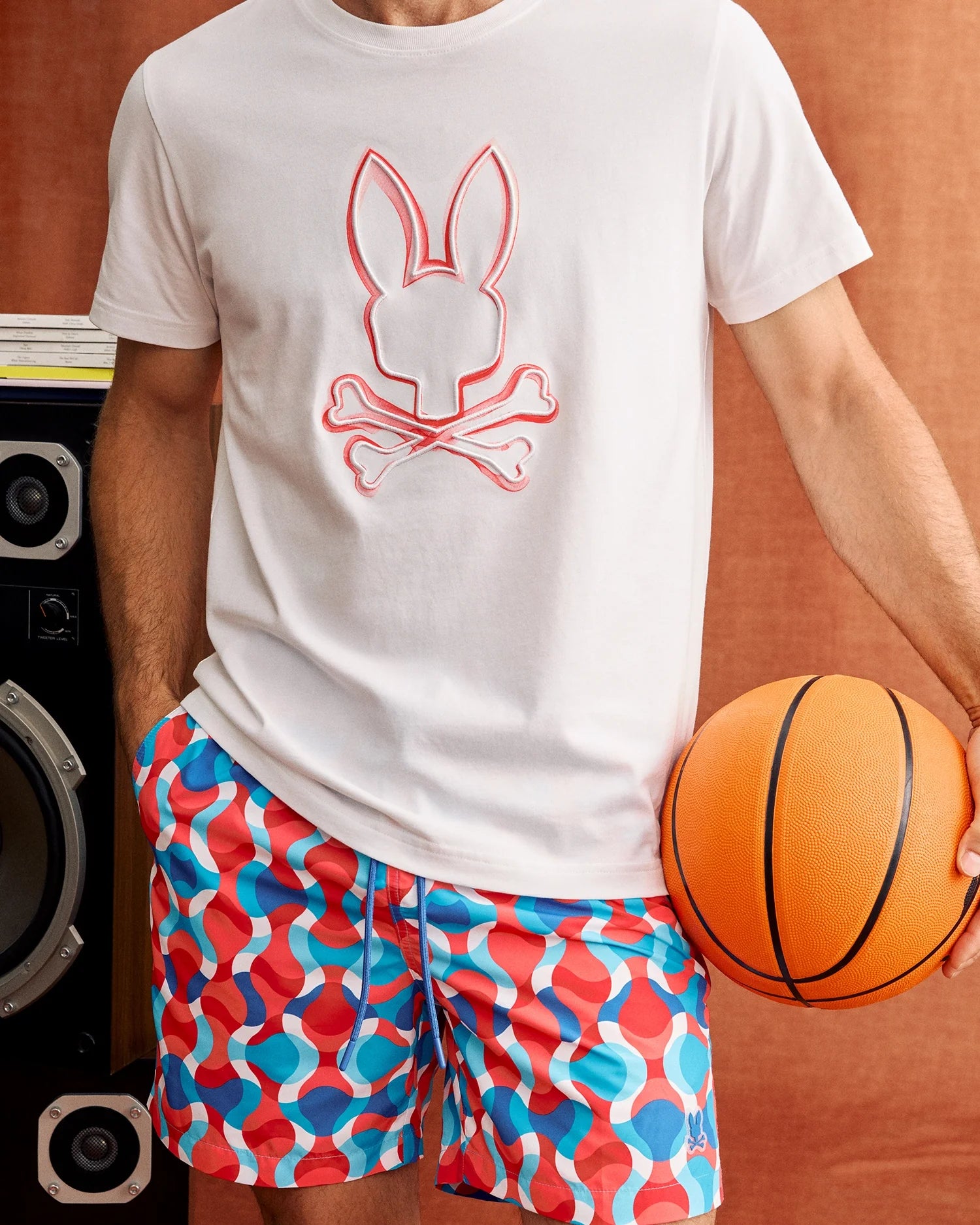 A person wearing a white t-shirt featuring a bunny and crossbones graphic and vibrant Psycho Bunny MENS WALTER LIGHTWEIGHT PRINT SWIM TRUNK (B6W863D200) stands holding an orange basketball. A large black speaker is visible in the background against a brownish backdrop.