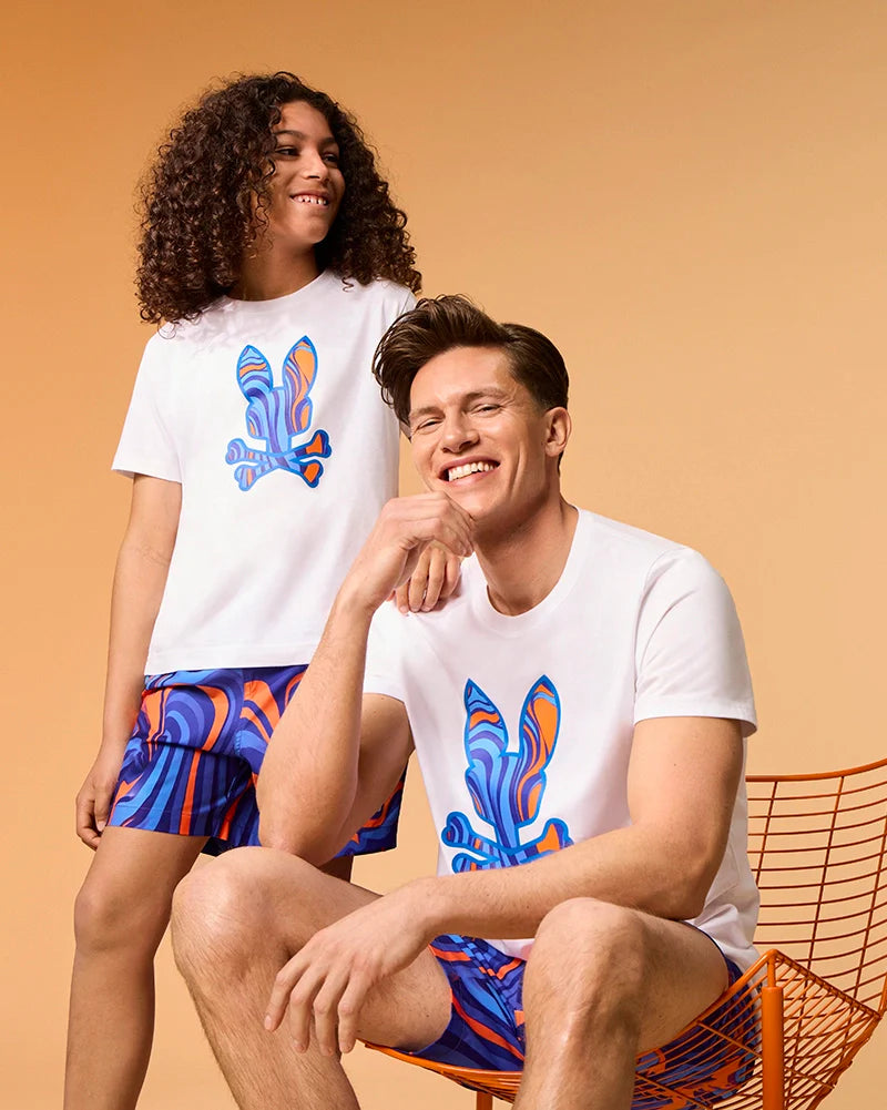 A young person with curly hair and an adult with straight hair are seen sitting and standing against a warm-toned background. Both are dressed in matching outfits, including white T-shirts adorned with colorful abstract rabbit designs, paired with the Psycho Bunny KIDS NEVADA ALL OVER PRINT SWIM TRUNKS (B0W526C200). These multicolored trunks feature quick-dry fabric in blue, orange, and purple patterns.