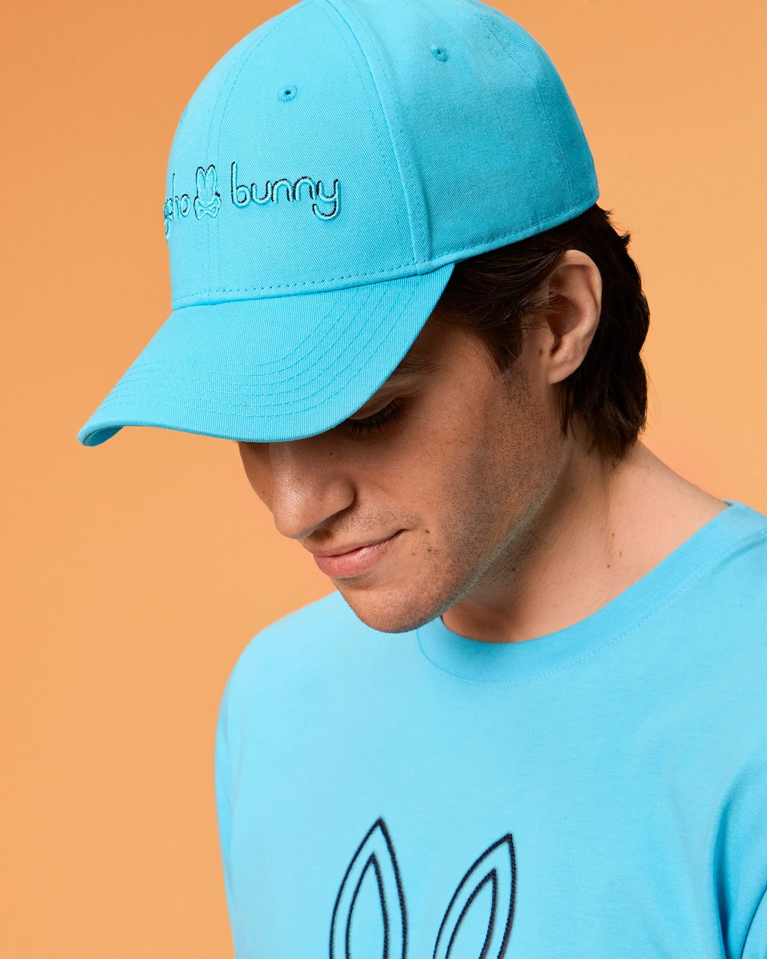 A person wearing a light blue Psycho Bunny MENS LOUISE BASEBALL CAP and matching t-shirt, both featuring an embroidered bunny logo. The background is a warm beige color. The person is looking down, and their brown hair peeks out from under the cap, which has the text "Psycho Bunny" on it.