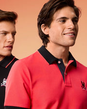 Two men are depicted in the image with a gradient orange background. The man in the foreground is wearing a red MENS KAYDEN PIQUE POLO - B6K675C200 by Psycho Bunny with a black collar featuring a small skull and crossbones emblem. The man behind him sports a black shirt with contrasting stripes on the red-trimmed collar.