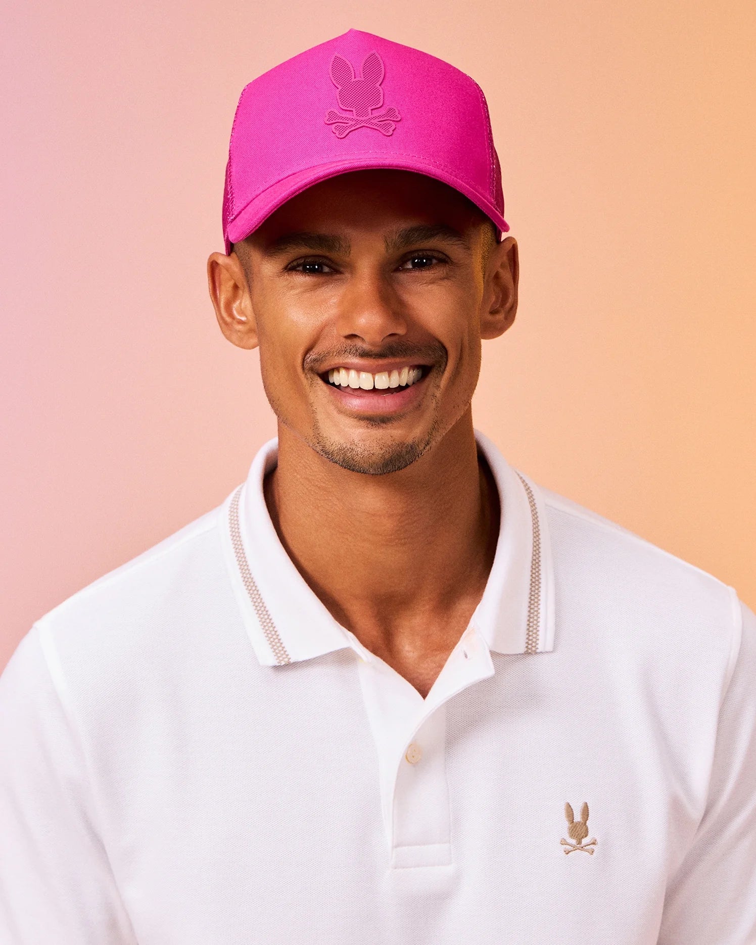 A smiling person wearing a bright pink Psycho Bunny MENS RIVIERA TRUCKER CAP - B6A667C200 with a logo of a bunny and crossbones, and a white polo shirt with a similar bunny logo evokes early 2000s nostalgia. The background showcases a soft gradient of peach and light pink.