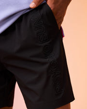 Close-up of a person wearing black MENS MELVIN SWIM TRUNK - B6W665C200 with the brand name "Psycho Bunny" embossed vertically along the left thigh. The person has their hand in the pocket of the quick-dry swim trunk. The background is a soft peach color.