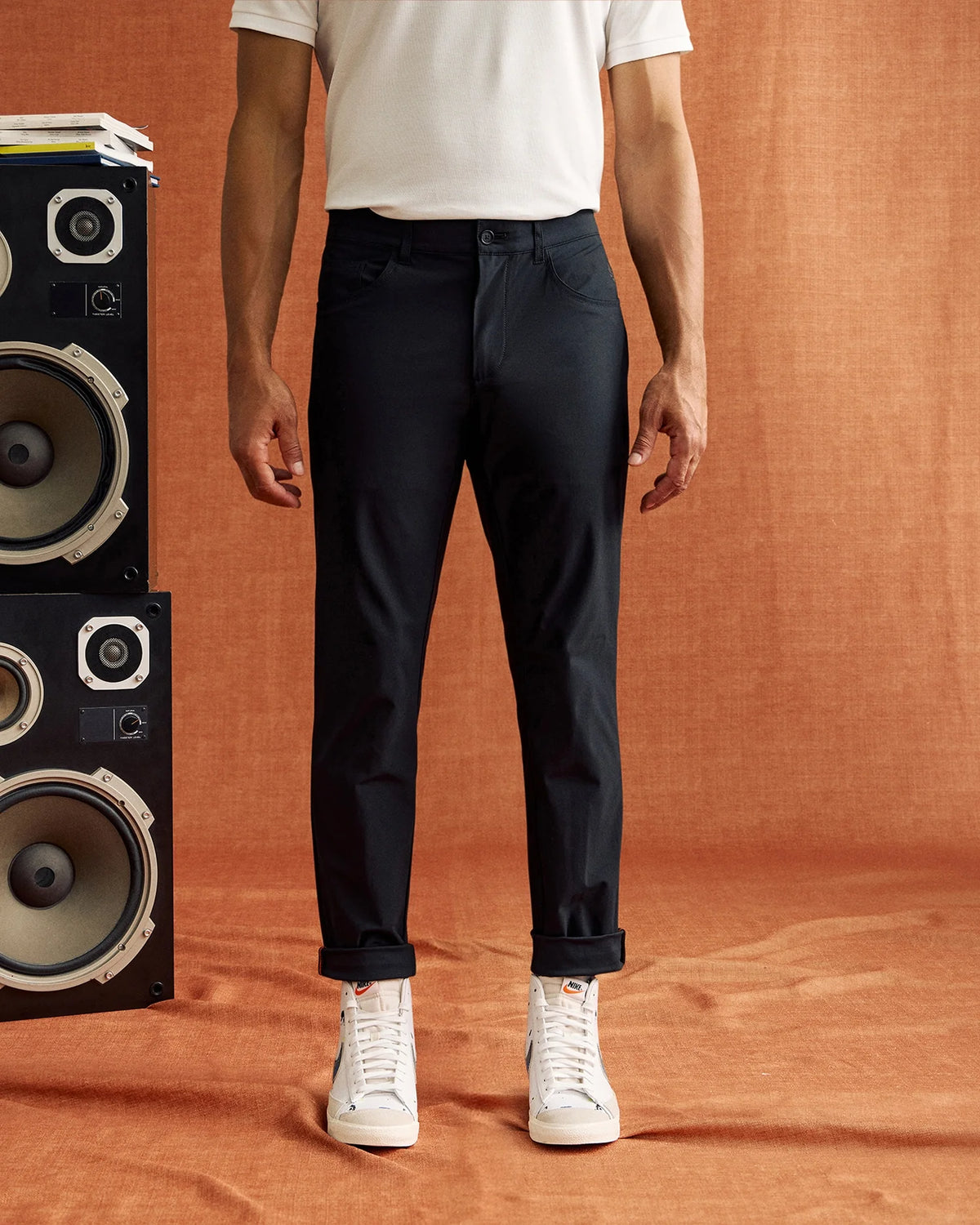 A person wearing Psycho Bunny's MENS GABLE REGULAR FIT SPORT PANT (model B6P760ARCN) made of stretch cotton-nylon textile and white sneakers stands against a burnt orange fabric backdrop, with only the lower half of their body visible. To the left, part of a black speaker with multiple audio drivers is in view.