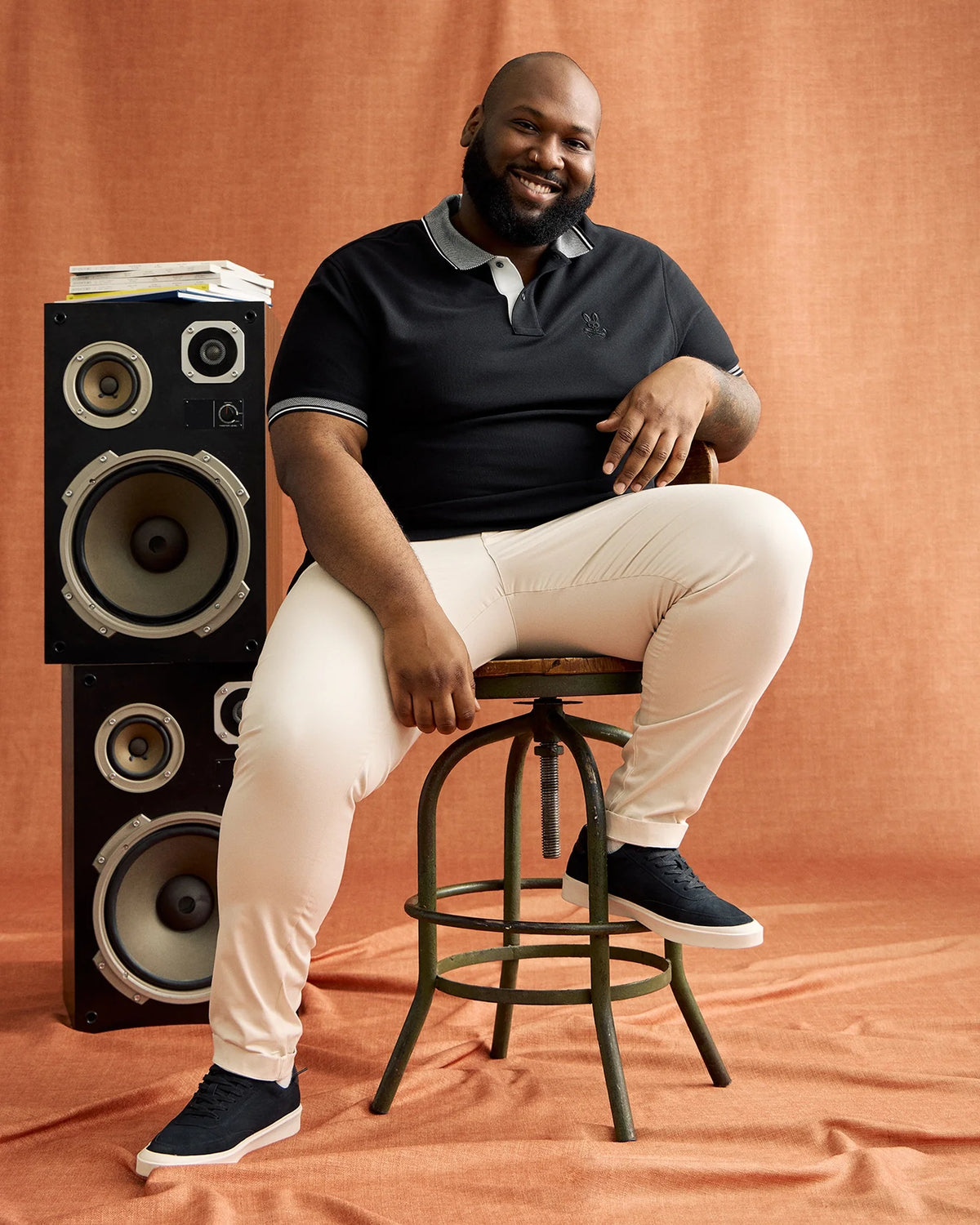 A man sits on a wooden stool, smiling, with one hand on his knee. He's wearing a black Psycho Bunny BIG AND TALL MALTON PIQUE POLO SHIRT made of premium Pima cotton, light pants, and dark shoes. Large speakers with papers are beside him against an orange curtain backdrop.