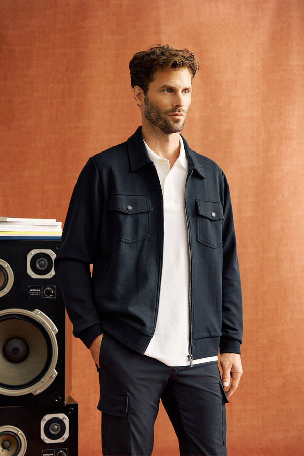 A man with a beard stands in front of a fabric backdrop, wearing the sleek black MENS OROVILLE BOMBER JACKET - B6J712A200 by Psycho Bunny over a white shirt and black pants, with a large speaker partially visible beside him.