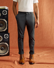 A person dressed in Psycho Bunny's MENS GILMAN CHINO PANT in gray, paired with a white shirt and brown leather boots, stands beside a stack of large speakers. The scene is set against a backdrop of warm orange-brown fabric. With hands tucked into their pockets, the ensemble radiates both style and comfort.