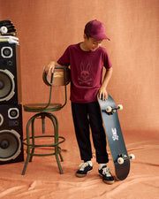 A boy in a maroon Psycho Bunny KIDS MALTON TRUCKER CAP, rabbit-logo T-shirt, black pants, and sneakers stands beside a branded skateboard. Embracing an active lifestyle, he leans on a wooden chair with two large speakers looming behind.