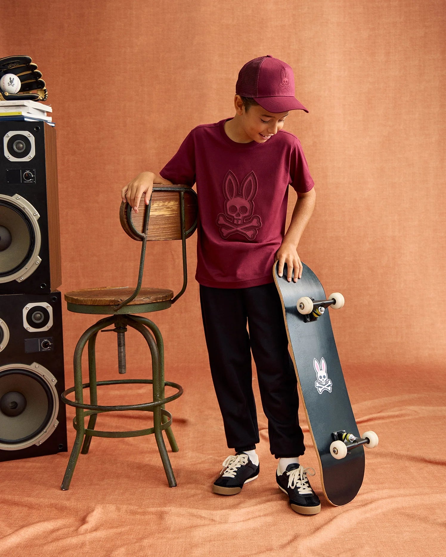 A boy in a maroon Psycho Bunny KIDS MALTON TRUCKER CAP, rabbit-logo T-shirt, black pants, and sneakers stands beside a branded skateboard. Embracing an active lifestyle, he leans on a wooden chair with two large speakers looming behind.