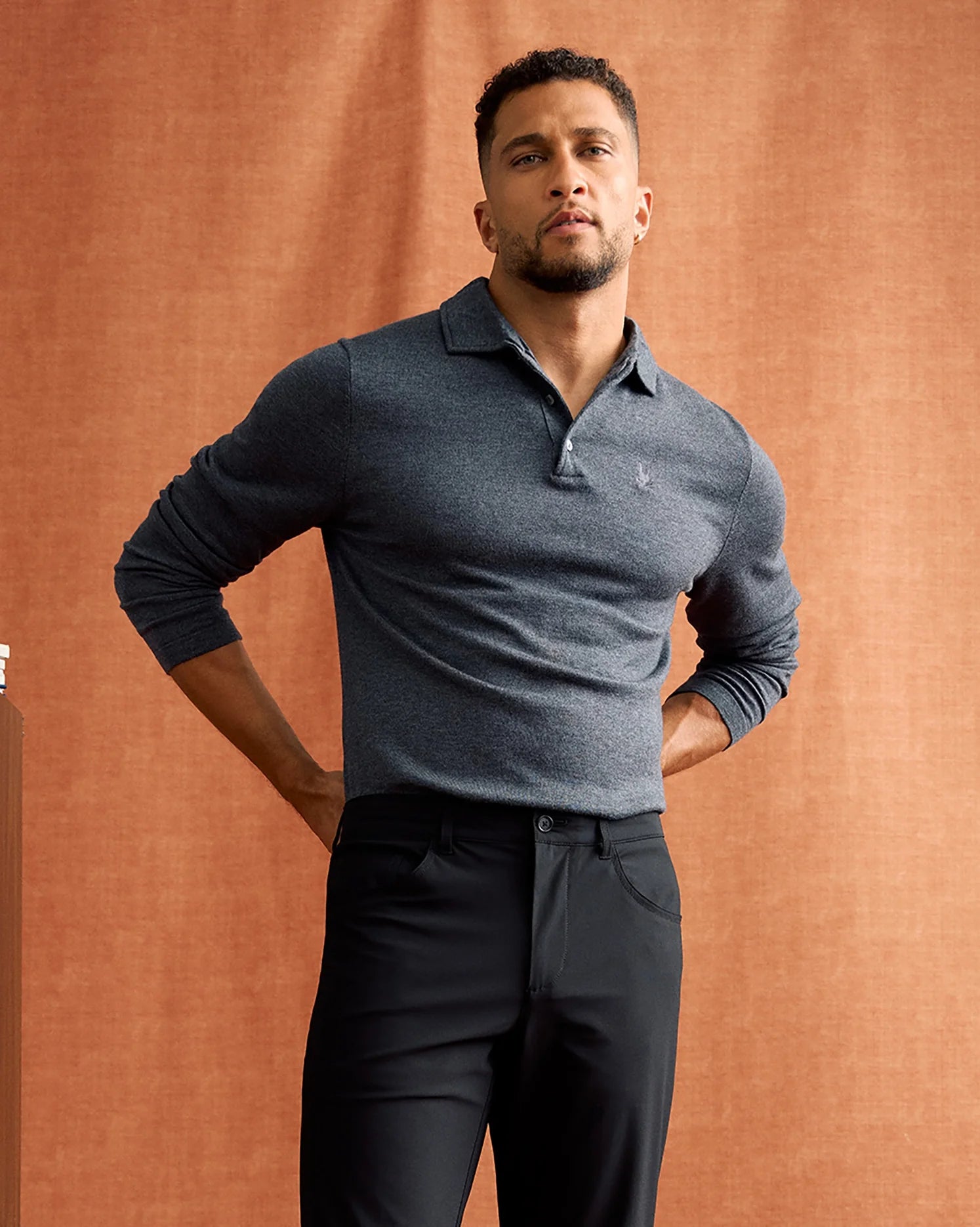 A man stands confidently against a warm, brown textured background, wearing the MENS BRADSHAW POLO SWEATER by Psycho Bunny in dark gray and paired with black pants, with his hands in his back pockets.