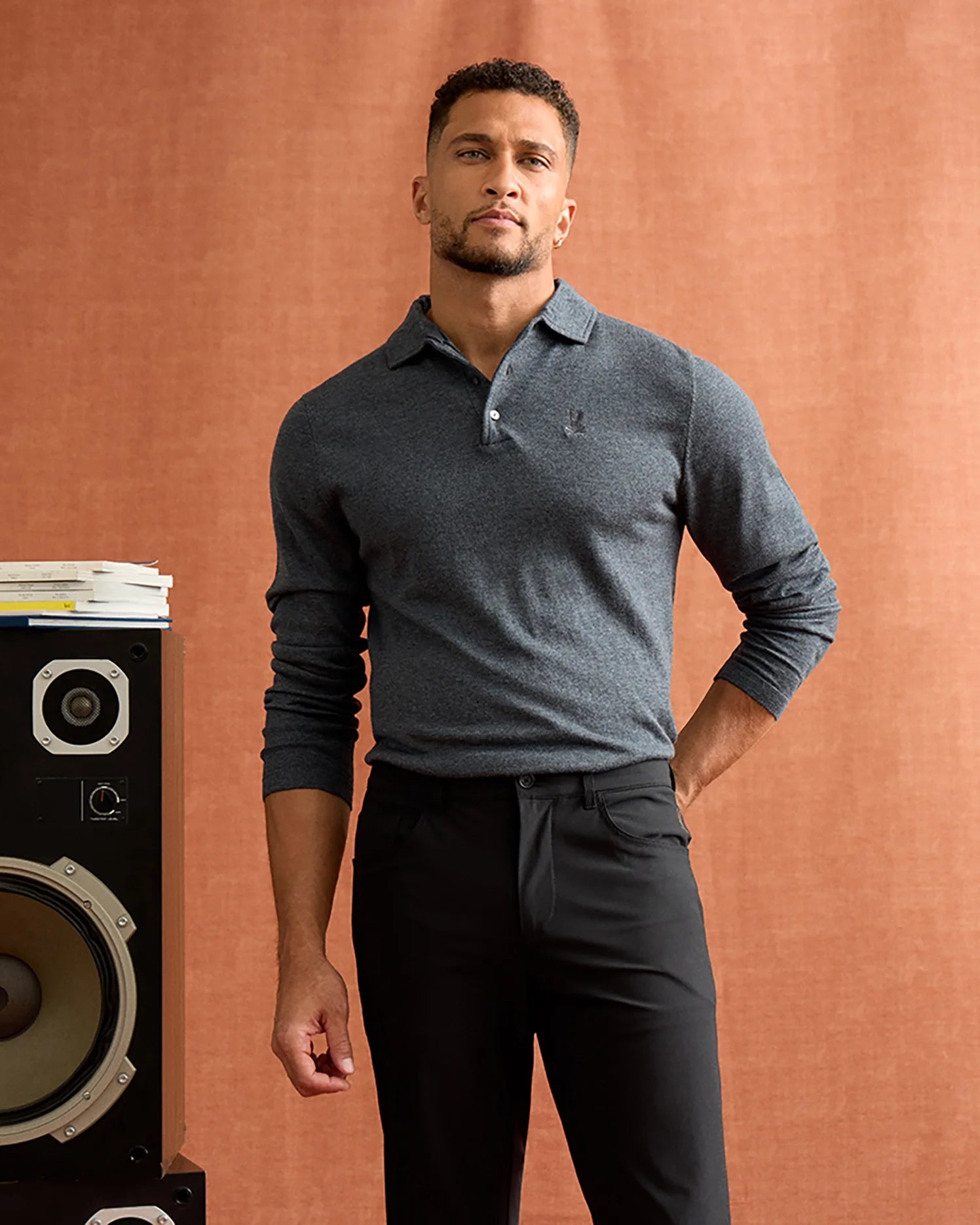 A man stands confidently in front of a brown backdrop, wearing the MENS BRADSHAW POLO SWEATER by Psycho Bunny and black pants. Next to him is a large speaker with a stack of books on top, contributing to an atmosphere of thoughtful luxury.
