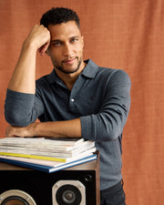 A man with short curly hair leans against a stack of books, resting his elbow on them. He is dressed in a Men's Bradshaw Polo Sweater by Psycho Bunny and stands beside a speaker. The rich texture of the warm brown fabric in the background complements his elegant style.