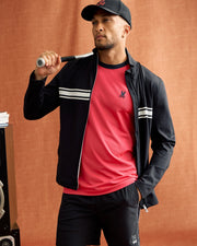 A person wearing a black cap and athletic outfit holds a baseball bat over their shoulder. They are wearing the MENS TERRELL SPORT ZIP UP JACKET by Psycho Bunny, which features white stripes, over a red shirt and black shorts. A speaker is partially visible in the background against a brown backdrop.