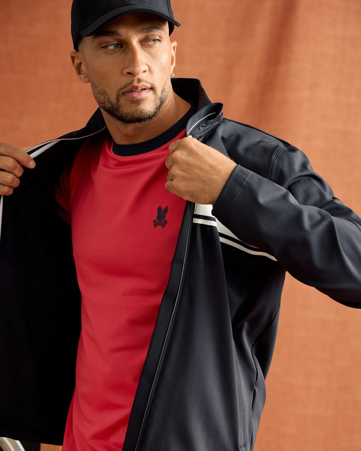 A person wearing the MENS TERRELL SPORT ZIP UP JACKET by Psycho Bunny and a black cap over a red shirt with a small emblem looks off to the side against a brown background, partially unzipping the jacket.