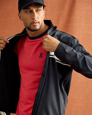 A person wearing the MENS TERRELL SPORT ZIP UP JACKET by Psycho Bunny and a black cap over a red shirt with a small emblem looks off to the side against a brown background, partially unzipping the jacket.