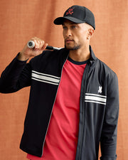 A man wearing the Psycho Bunny MENS TERRELL SPORT ZIP UP JACKET - B6J643C200, which boasts a black and red design with white stripes, holds a tennis racket over his shoulder. He complements the look with a black cap and a red shirt against the warm brown backdrop.